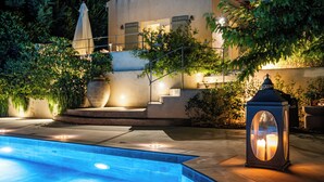 A swimming pool with a stone border, a lantern, and a staircase leading to a building with a lighted entrance. - Kalliroi - Exquisite Villa Designed Exclusively For Two with Large Private Pool. (Pylaros)