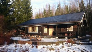 A wooden lodge with a covered patio and picnic tables, surrounded by snow and trees.