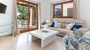 Ein modernes Wohnzimmer mit einer Couch, einem Couchtisch und einem Glastisch. Eine Schiebetür aus Glas führt zu einem Außenbereich mit Tisch und Stühlen.