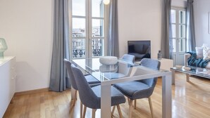 A modern living room with a glass dining table, grey chairs, a flat-screen TV, and large windows with grey curtains. - SibsBcn- Balmes - Romantic Central Cozy (Barcelona)