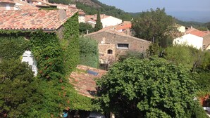 Un pueblo en una ladera con edificios de piedra tradicionales, algunos con tejados rojos, rodeado de abundante vegetación y colinas al fondo.