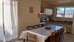 Un coin repas comprenant une table, des chaises, un panier de fruits, une cuisine avec des placards, une fenêtre et un mur recouvert de carreaux.
