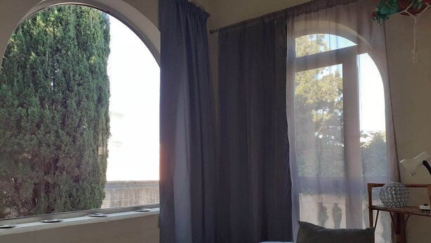 Une chambre à coucher avec une grande fenêtre en arc de cercle, un lit avec deux oreillers et une table de chevet agrémentée d’un objet décoratif.