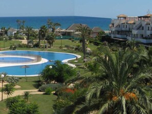 Een resort met een groot rond zwembad, palmbomen en uitzicht op zee.