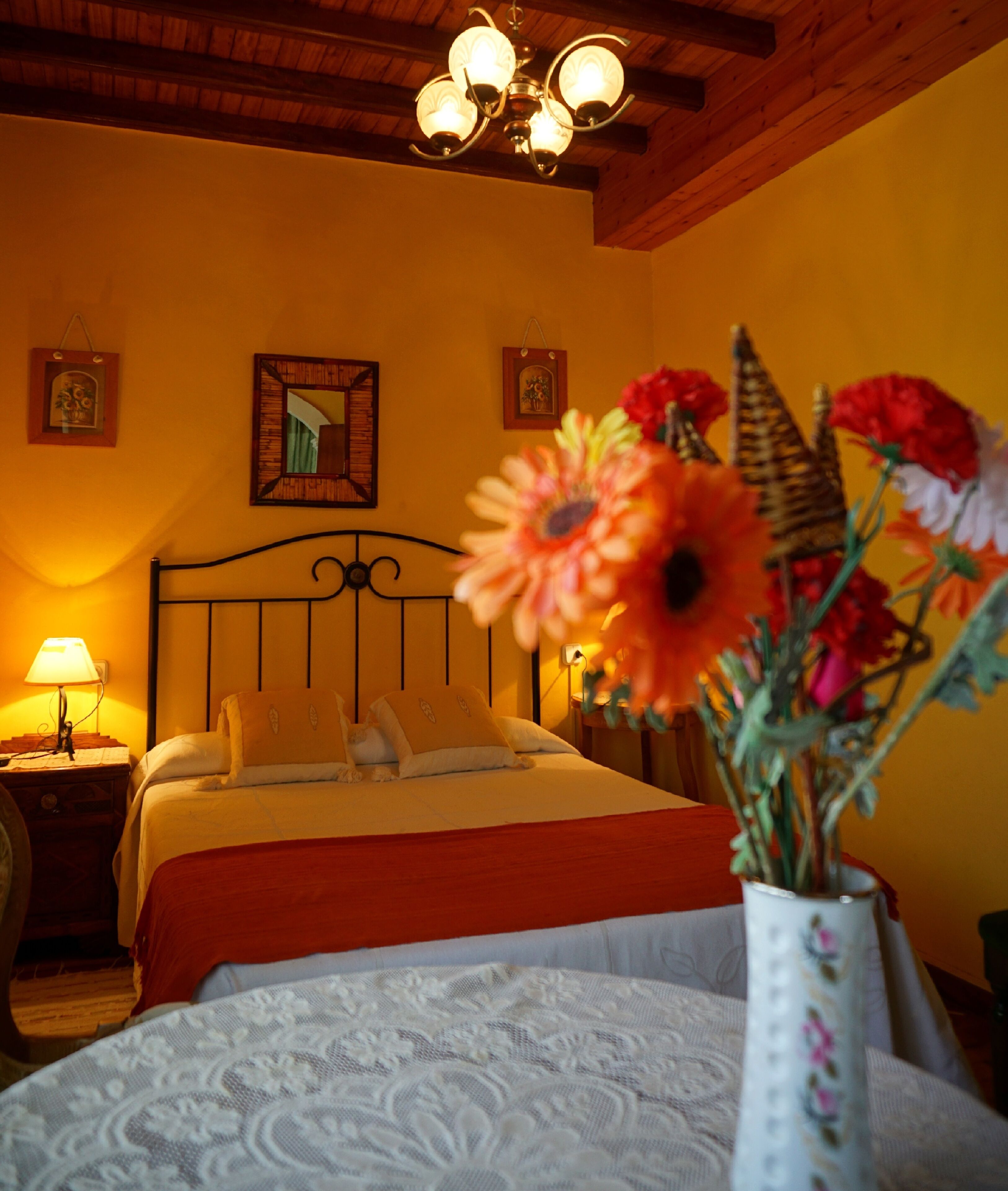 Une chambre à coucher avec un lit, une table de chevet, une lampe, un miroir et un vase avec des fleurs.