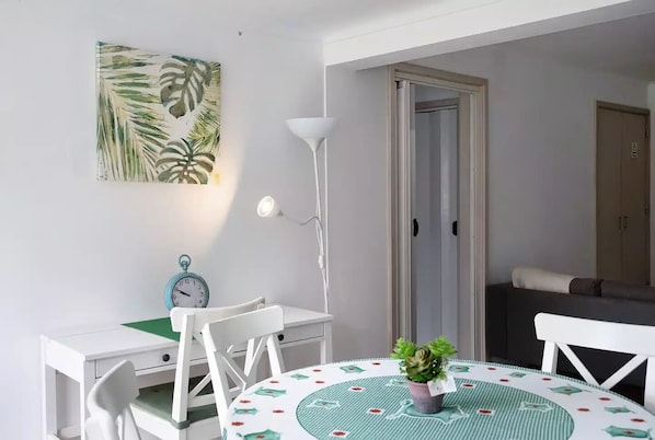 Une pièce avec une table ronde, des chaises, une horloge, une lampe et une porte qui donne sur une autre pièce.