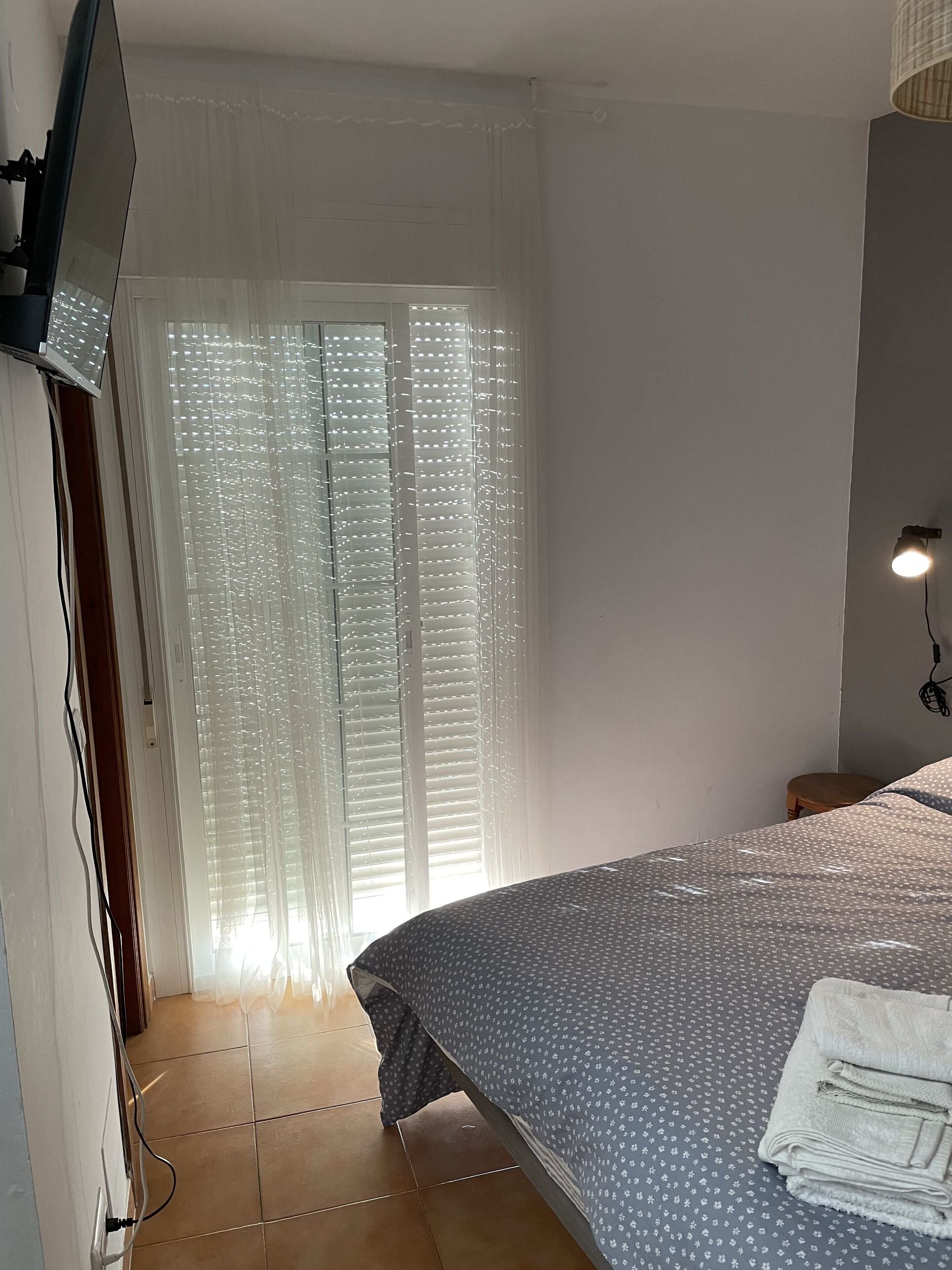 Ein Schlafzimmer mit Bett, Wand-TV, Fenster mit Jalousien und Nachttischlampe.