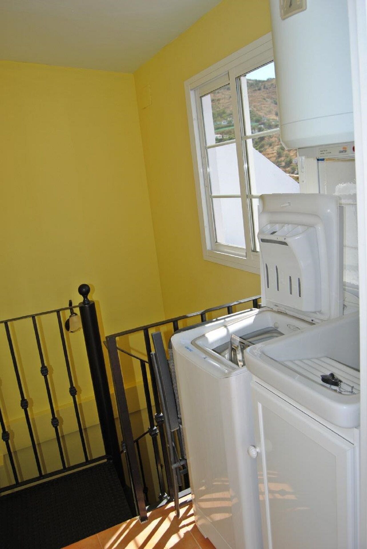 A laundry area with a washing machine and dryer, a window with a view of a hilly landscape, and a black metal railing.