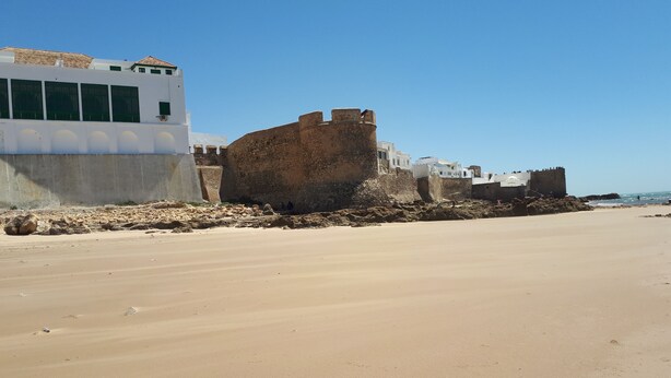 Ein sandiger Strand mit einer historischen Festung und einem weißen Gebäude mit grünen Fenstern.