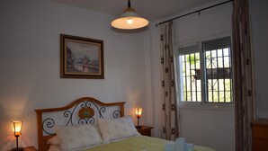 Un lit bien fait, recouvert d’une couvre-lit jaune, deux lampes de chevet, un tableau encadré au mur et une fenêtre avec des rideaux.