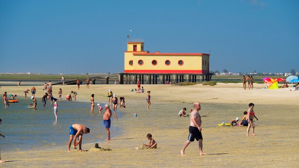 Ein Strand mit Menschen, die verschiedene Aktivitäten ausüben, ein Steg mit einem farbenfrohen Gebäude und strahlend blauem Himmel.