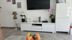 Una sala de estar moderna con un televisor de pantalla plana, mueble blanco de entretenimiento y elementos decorativos como flores y una estatua de Buda.