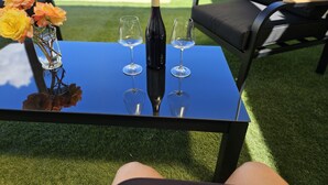 Una mesa junto a la piscina con una botella de vino y dos copas, rodeada de sillas y un florero con flores.