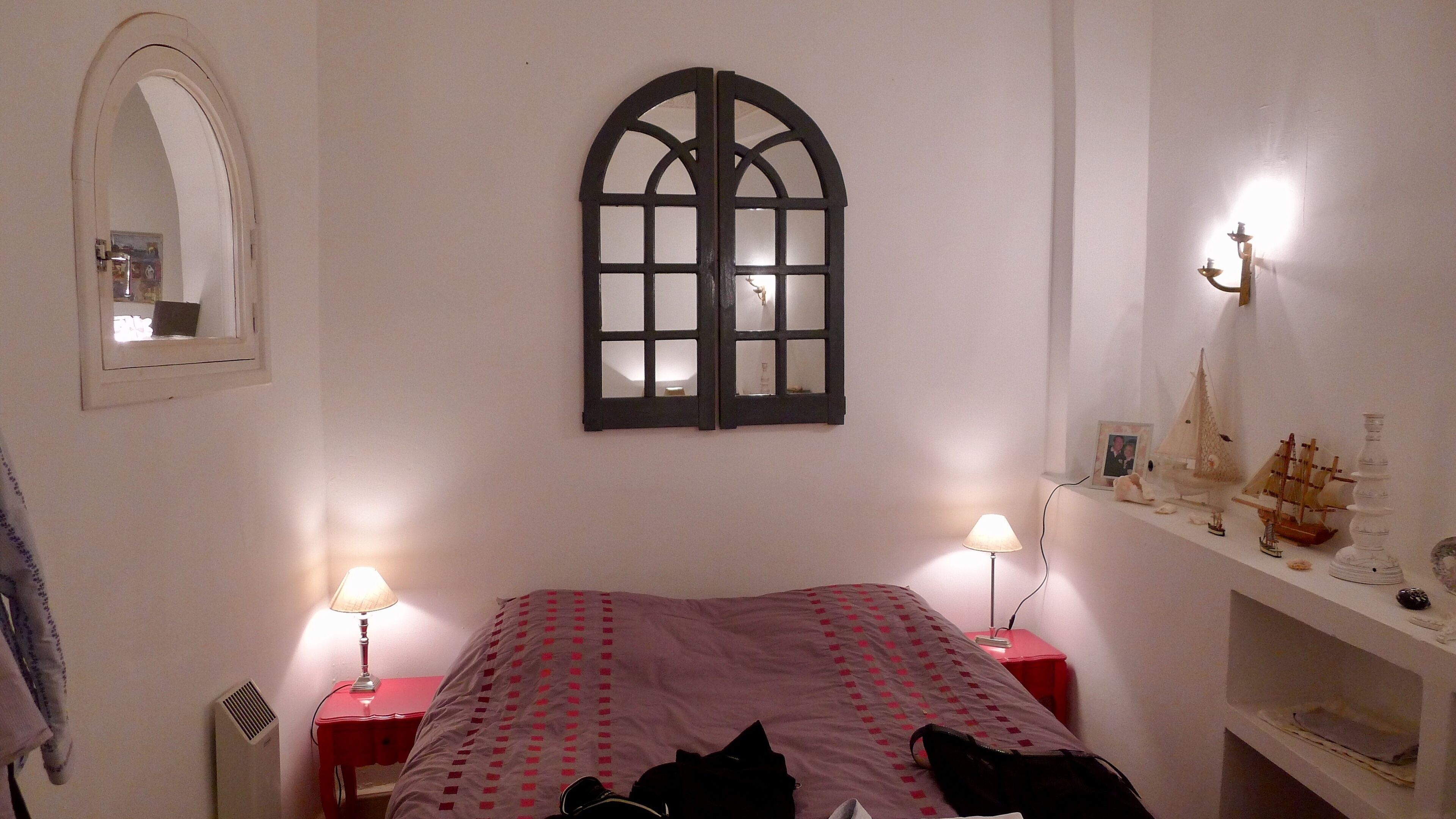 Un dormitorio con cama, dos lámparas de noche, un espejo, una ventana con marco decorativo y una estantería con barcos modelo.