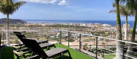 A balcony with green artificial turf, two lounge chairs, and a view of a coastal cityscape.