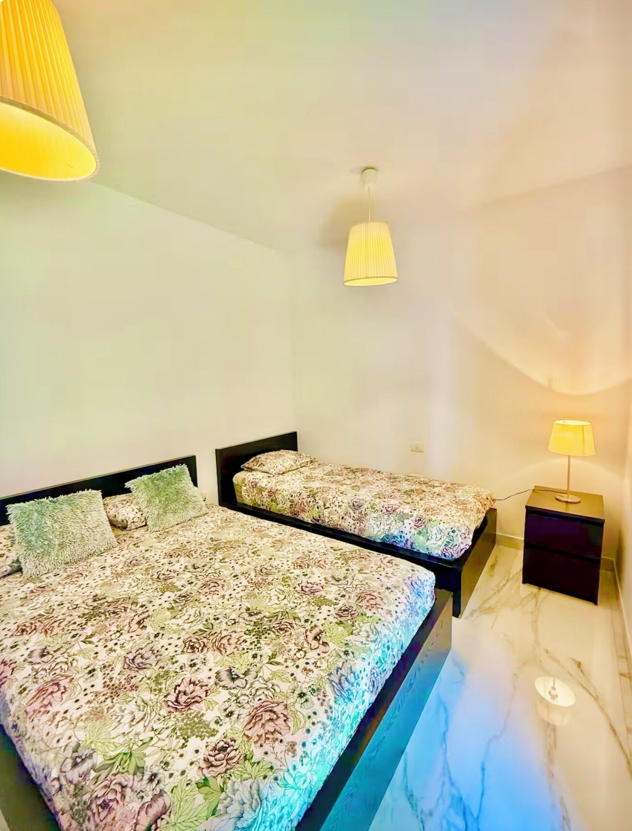 A bedroom with two beds, each with floral bedding, a nightstand with a lamp, and a hanging lamp.