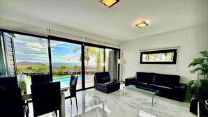 Un salón espacioso con un amplio ventanal que da a una piscina, un sofá de cuero negro, una mesa de centro de vidrio y una zona de comedor con sillas.