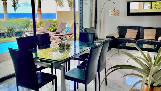Una sala moderna con una mesa y sillas de vidrio, un sofá negro y vista al océano.