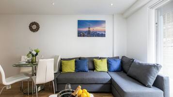 A modern living room with a grey sectional sofa, a dining table with chairs, a small side table with a vase of flowers, and a cityscape painting on the wall.