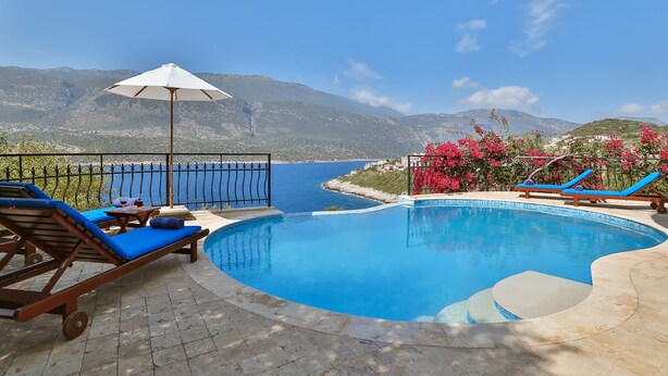Un espace aménagé au bord de la piscine, avec des chaises longues et une table, offrant une vue sur un plan d’eau et des montagnes.