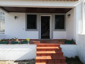 Una casa con un porche de baldosas, dos ventanas con contraventanas y plantas en macetas en los escalones.