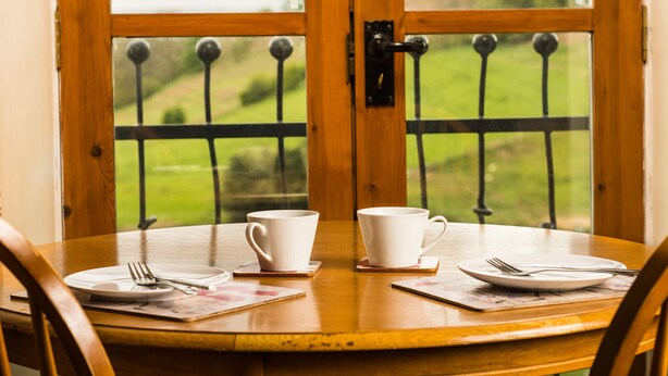A dining table with two white cups and plates, set with cutlery, in front of a wooden door with a metal gate.
