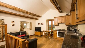 A cozy living room with a sofa and a TV, wooden beams on the ceiling, a dining area with a table and chairs, and a kitchen with wooden cabinets and appliances.