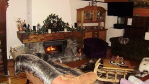 Una sala de estar acogedora con chimenea de piedra, un fuste de madera y un árbol de Navidad. Hay un sillón mullido, una mesa de centro de madera y una mesita con un objeto decorativo.
