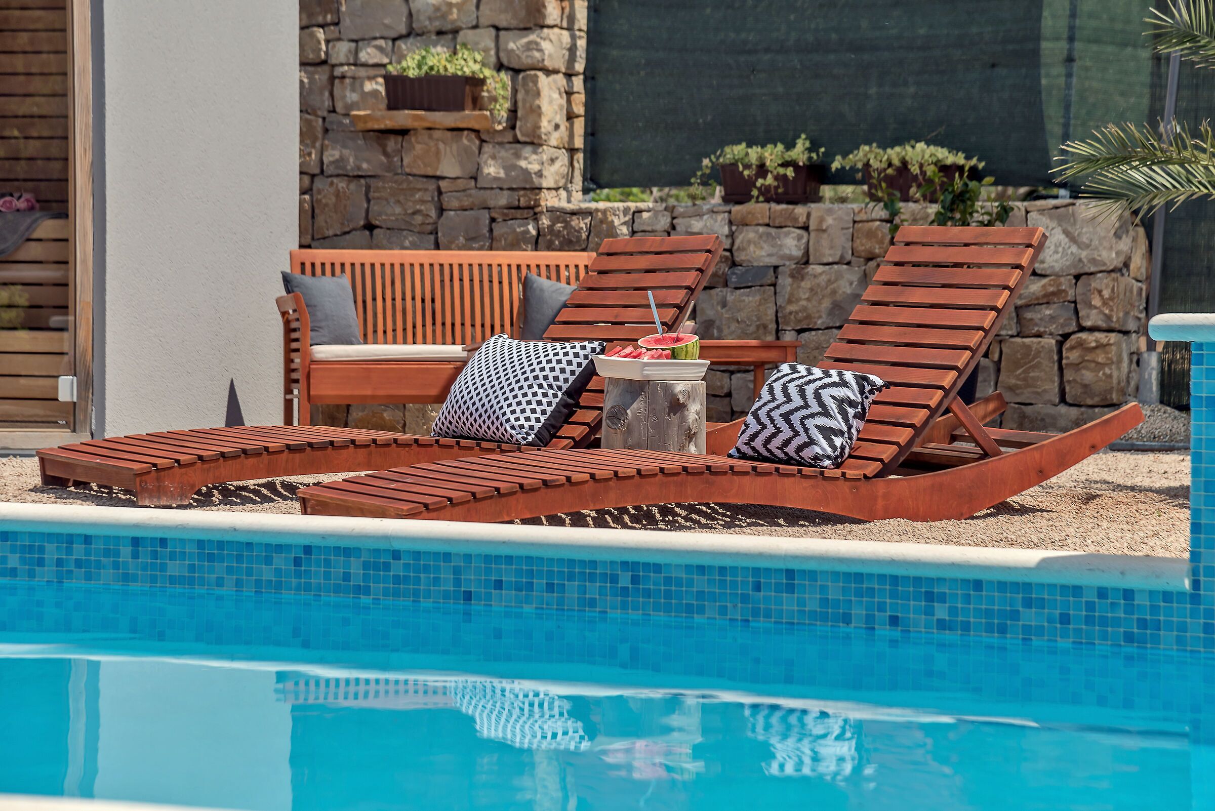 A poolside area with wooden lounge chairs and a table with a watermelon and drink.