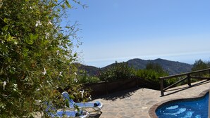 Une piscine en pierre avec une terrasse en bois, entourée d’une végétation luxuriante et offrant une vue sur les montagnes lointaines.