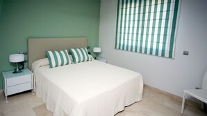 A bedroom with a bed, two bedside tables, a nightstand, a window with blinds, and a chair.