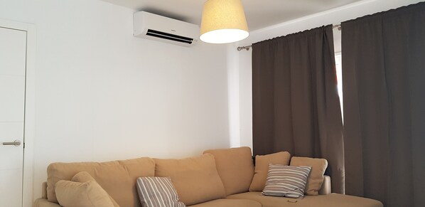 Ett vardagsrum med en beige soffa, en randig kudde, en takfläkt och ett fönster med mörka gardiner.