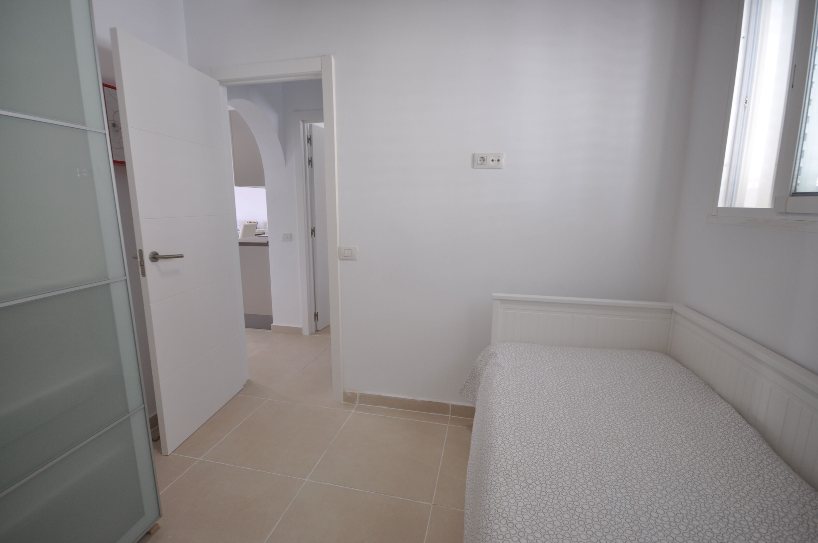 A small, white room with a bed, a door leading to another room, and a window.