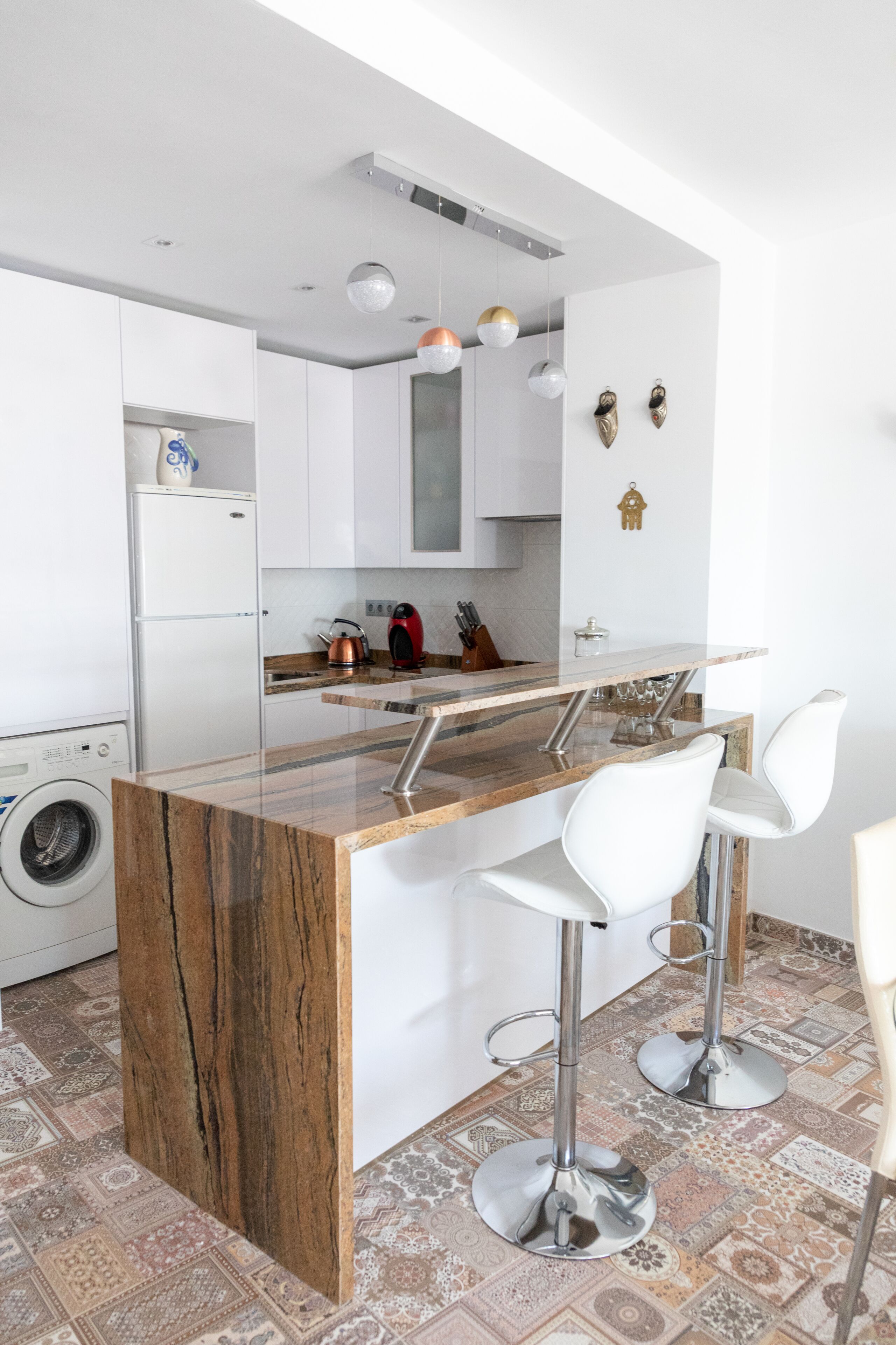 A modern kitchen with a marble island, white bar stools, and a washing machine in the corner.