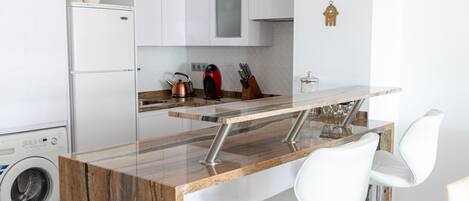 A modern kitchen with a marble island, white bar stools, and a washing machine in the corner.