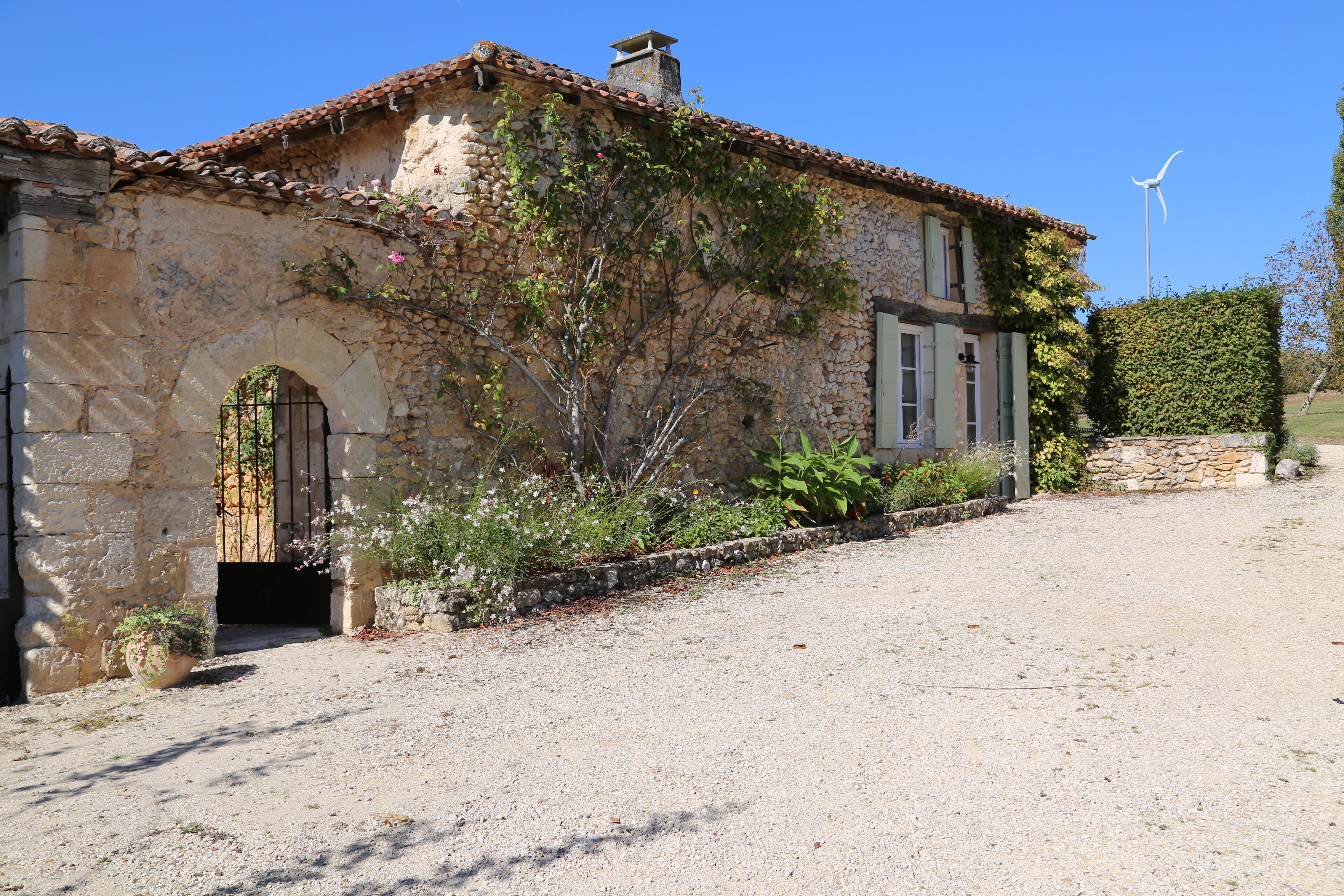 Un bâtiment rustique en pierre, avec une allée de gravier, une entrée en arc de cercle et une éolienne en arrière-plan.