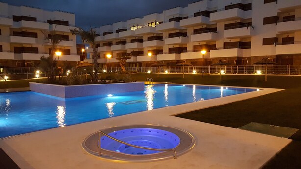 Una piscina con una vasca idromassaggio in primo piano, circondata da un edificio con numerosi balconi e illuminazione esterna.