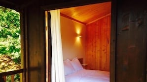 Una habitación de cabaña de madera con una cama, una ventana con cortinas y una lámpara fijada a la pared.