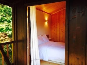 A wooden cabin room with a bed, a window with curtains, and a wall-mounted light.
