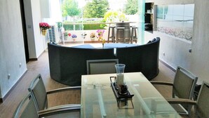 Uma sala de jantar moderna com mesa de vidro, cadeiras de metal, sofá preto e vista para o jardim através de uma porta de vidro.