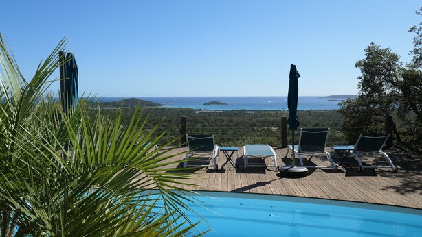 A poolside area with lounge chairs and an umbrella, overlooking a scenic view of the sea and hills.