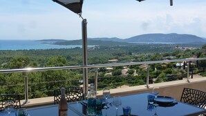 A dining table set with wine glasses, plates, and a bottle, overlooking a scenic view of the sea and mountains.