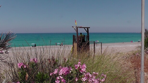 Une plage avec une tour de surveillance pour les sauveteurs, une eau turquoise et limpide, et quelques personnes qui se prélassent au soleil.