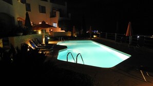 Une piscine nocturne, dotée de chaises longues et de parasols, entourée d’un bâtiment aux nombreuses fenêtres et d’un mur de briques.