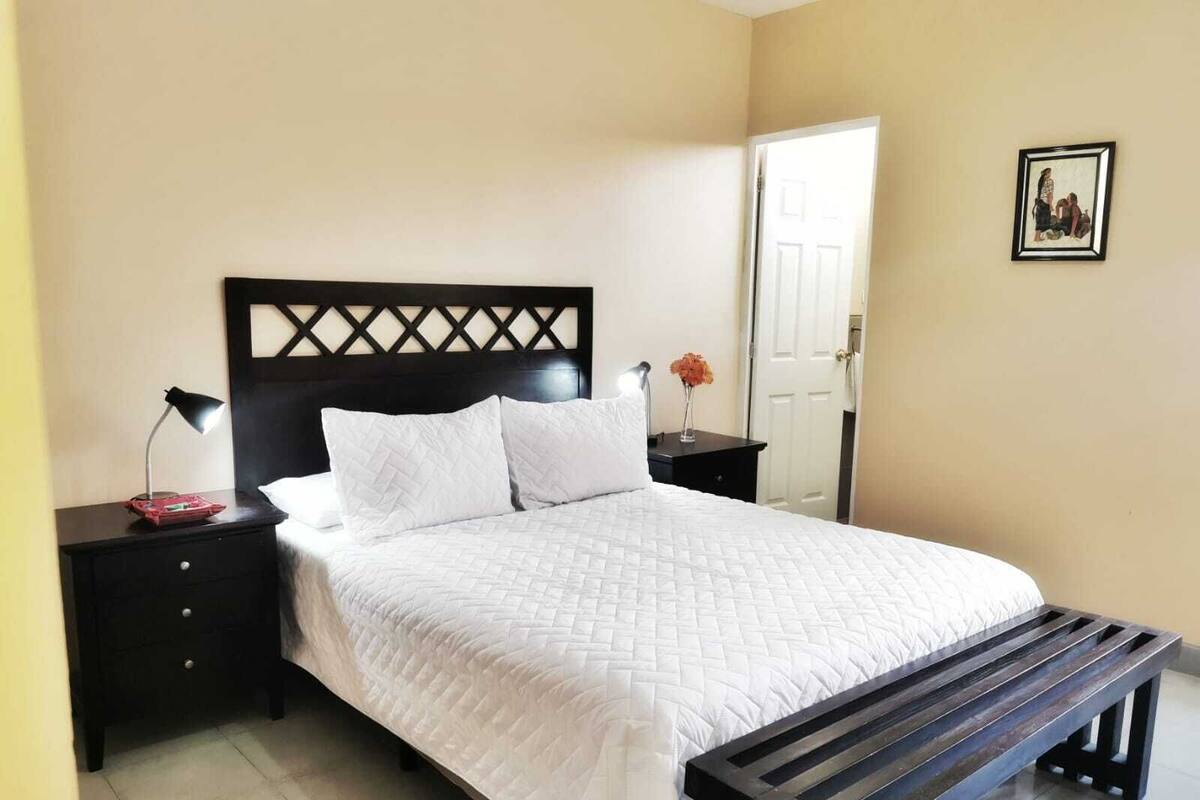 A neatly made bed with a wooden headboard, a bedside table with a lamp, and a bench at the foot of the bed.