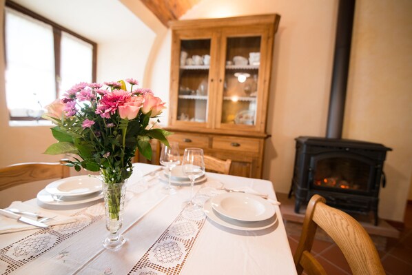 Una mesa de comedor dispuesta con platos, vasos y un jarrón de flores. Se observa un aparador de madera y una chimenea al fondo.