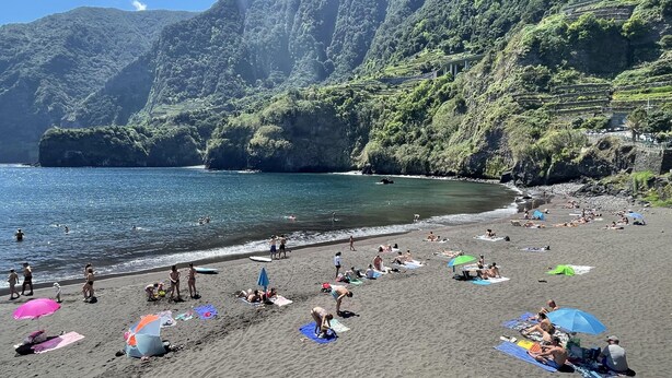 Une plage où des gens se prélassent au soleil et se baignent, entourée de collines verdoyantes.