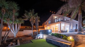 Une maison moderne à deux étages, dotée d’un balcon, d’un espace extérieur bien éclairé avec une pelouse artificielle et de palmiers.