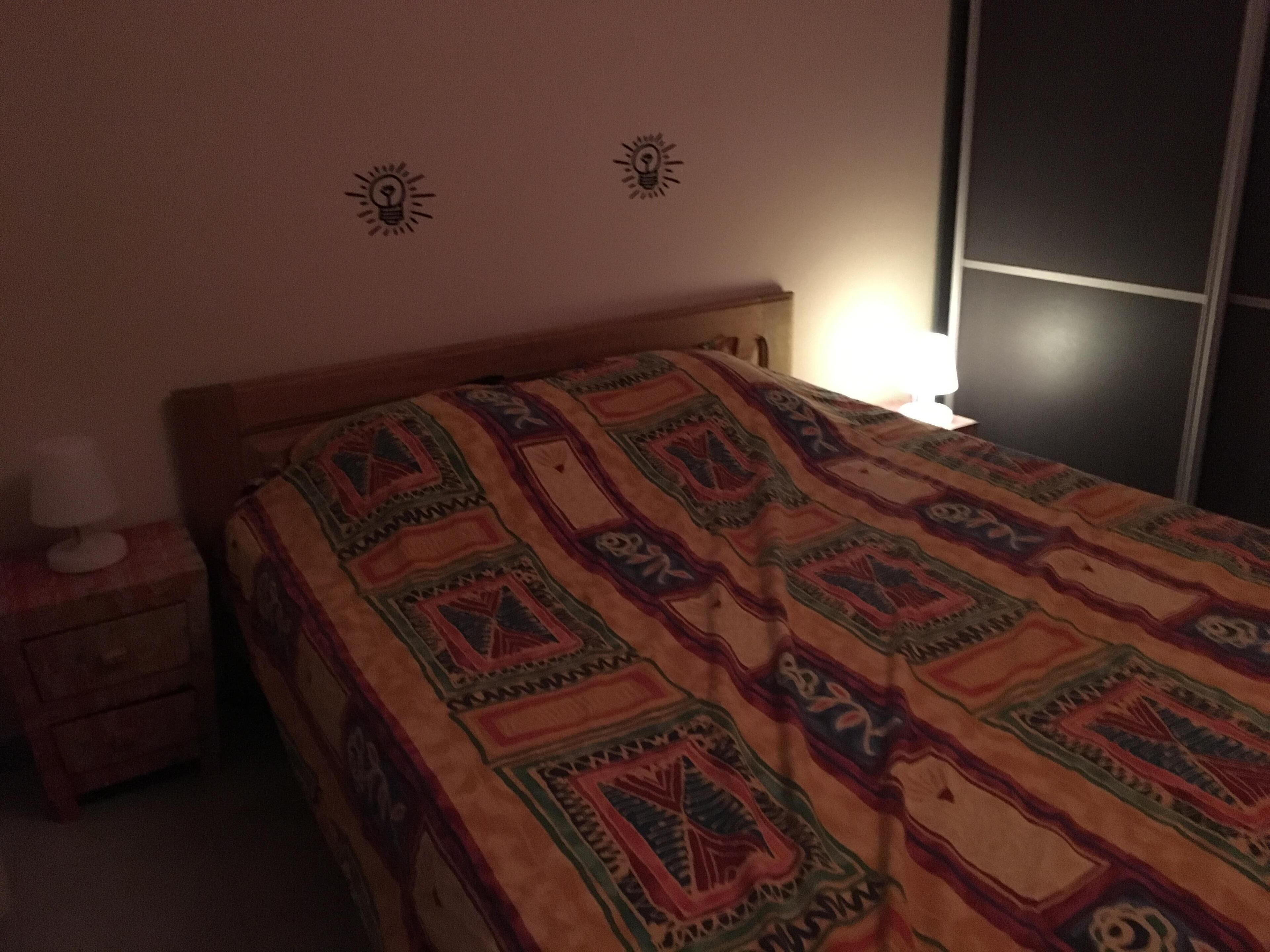 A bedroom with a bed, a nightstand, a lamp, and a wall with two light bulb icons.