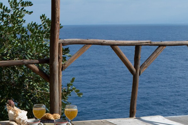 A wooden deck overlooking the sea with a rustic railing, a table with a shell, two glasses of orange juice, and a croissant.